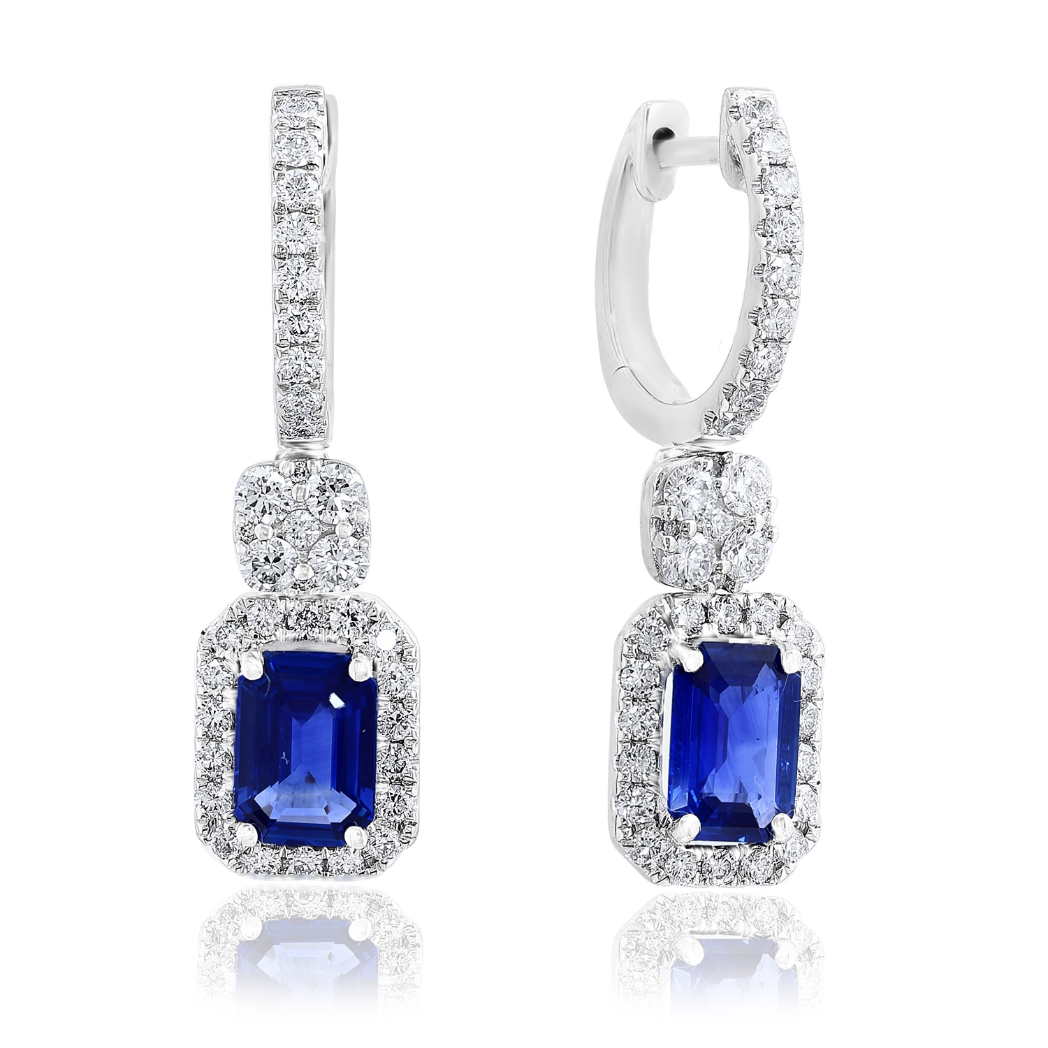 A pair of silver huggie hoop drop earrings featuring a rectangular emerald-cut blue sapphire center stone surrounded by a halo of small clear stones, with a smaller round blue stone above, set against a white background with a soft reflection.